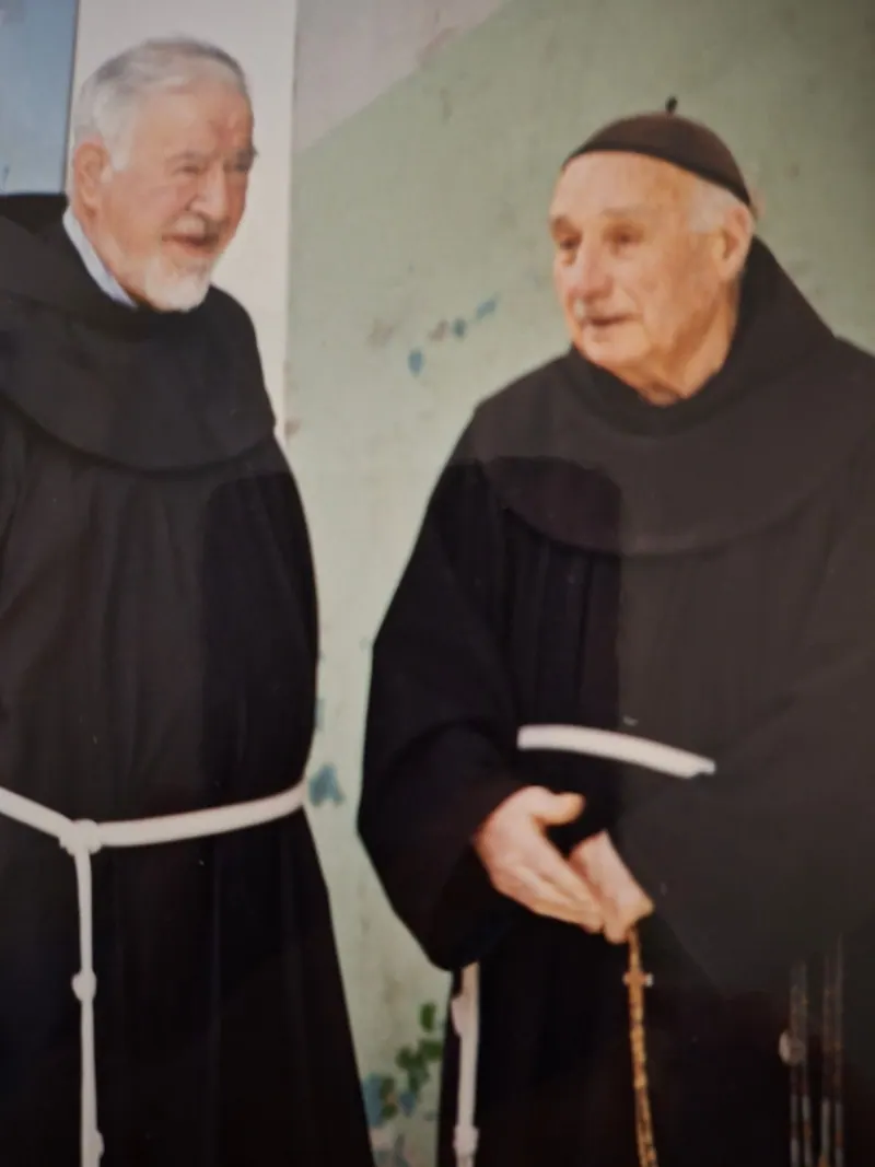 שני נזירים פרנציסקנים קשישים בגלימות חומות כהות וחבל לבן למותניהם משוחחים ליד קיר בתהלוכת יום שישי בדרך הייסורים; אחד מהם מחזיק מחרוזת תפילה.