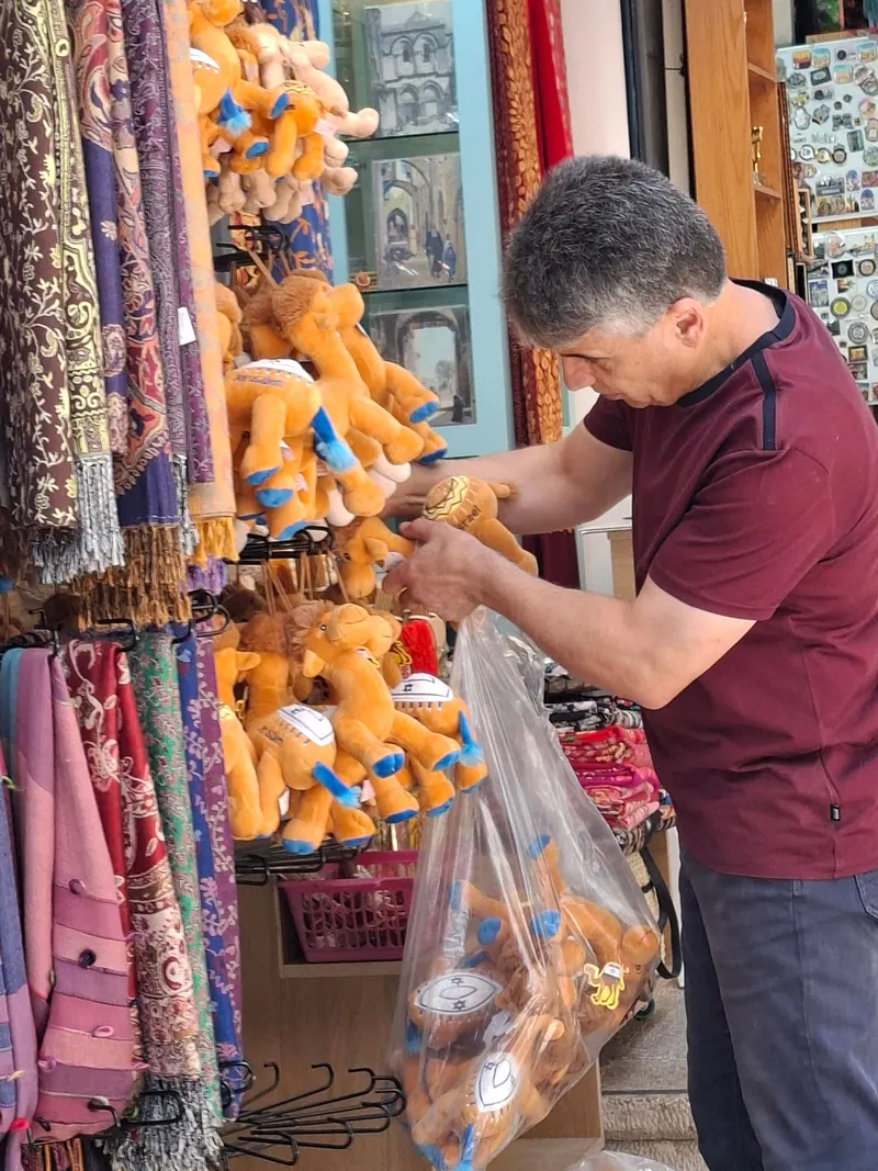 מוכר אורז גמלי פרווה בשקית ניילון בחנות מזכרות, לצד צעיפים תלויים.