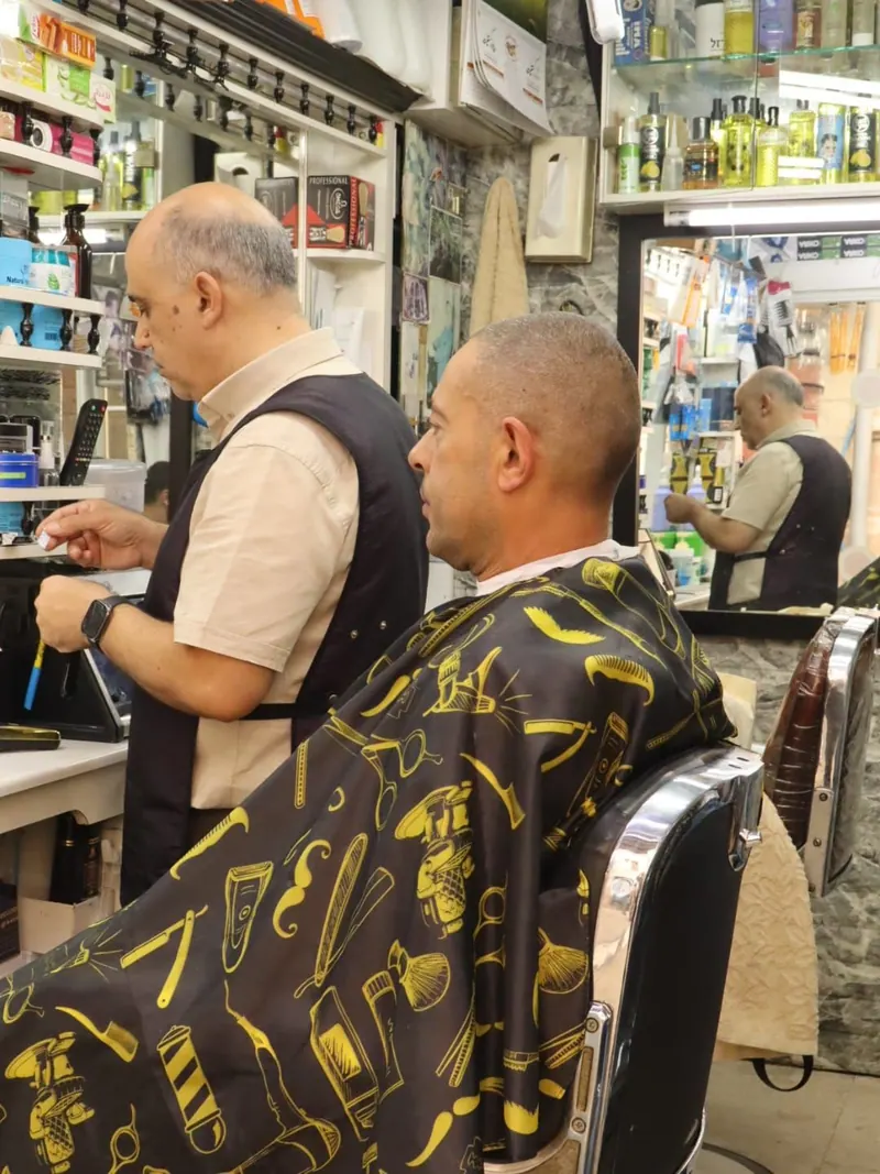 מספרה: לקוח יושב בכיסא עטוף בגלימה שחורה מעוטרת בכלי ספרות צהובים, והספר עומד לידו ליד הדלפק.