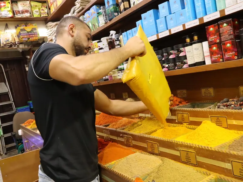 בחנות תבלינים: מוכר שופך כורכום משקית צהובה אל תאי תבלינים טחונים, ומאחוריו מדפי מוצרים.