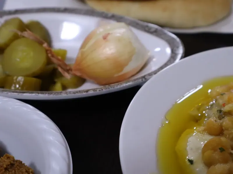 צלחת חומוס עם גרגרי חומוס ושמן זית, ולצדה מלפפונים חמוצים, בצל שלם ופיתה.