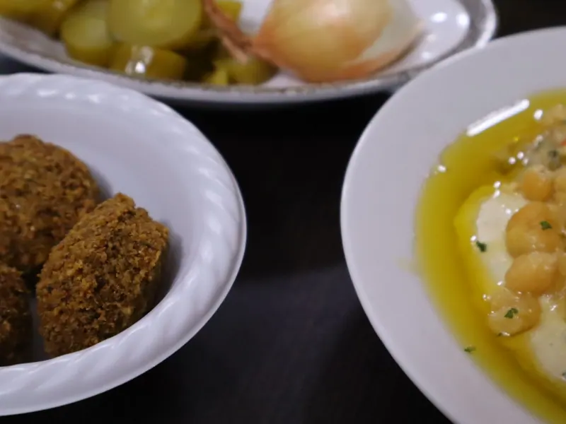 כדורי פלאפל בקערה לבנה לצד צלחת חומוס עם גרגרי חומוס ושמן זית; ברקע מלפפונים חמוצים ובצל.