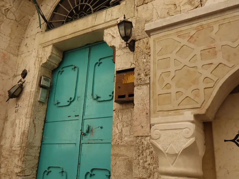 דלת מתכת בצבע טורקיז בסמטת אבן, עם פנס קיר ותיבת דואר, לצד קשת אבן מגולפת בדוגמה גאומטרית.
