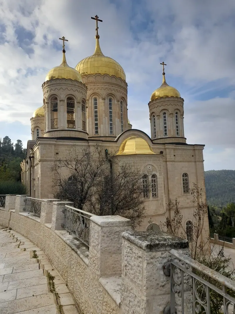 כנסייה לבנה עם כיפות בצל מוזהבות על ראש גבעה מיוערת, ולפניה מעקה אבן.
