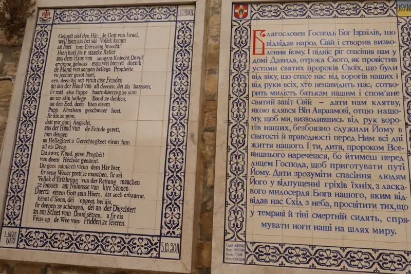 שני לוחות אריחים זה לצד זה עם תפילת Benedictus בלוקסמבורגית ובאוקראינית, במסגרות עיטורים כחולות.