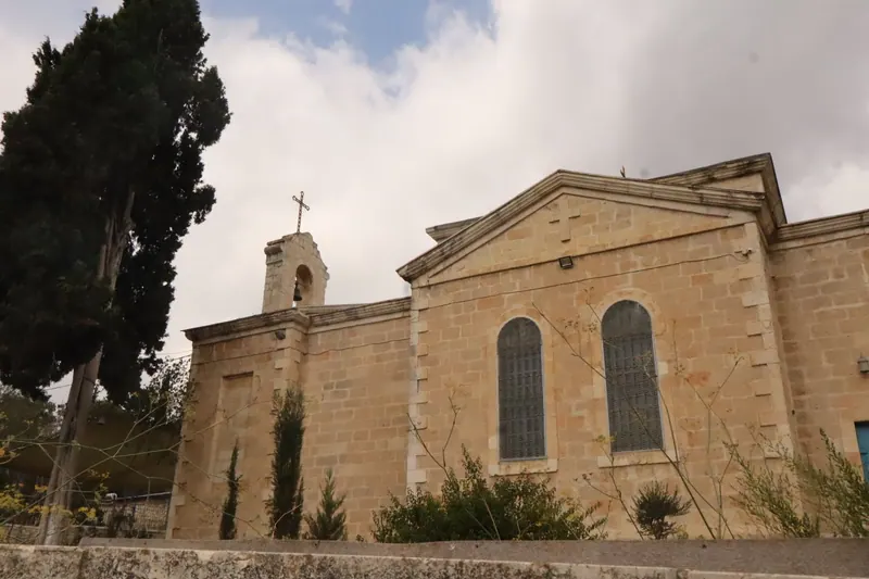 כנסיית אבן עם גמלון, שני חלונות קשתיים מסורגים ומגדלון פעמון עם צלב, לצד ברוש גבוה תחת שמיים מעוננים.