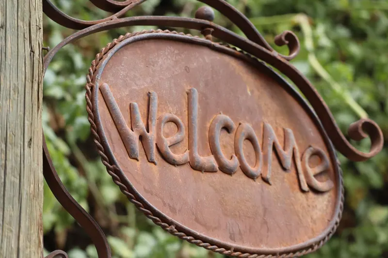שלט ברזל חלוד ועליו "Welcome" באותיות בולטות, תלוי על עמוד עץ מול עלווה.