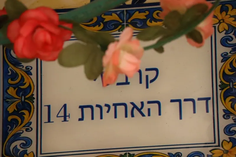 שלט קרמיקה במסגרת מעוטרת כחול וצהוב, ועליו "דרך האחיות 14"; ורדים מלאכותיים תלויים לפניו.