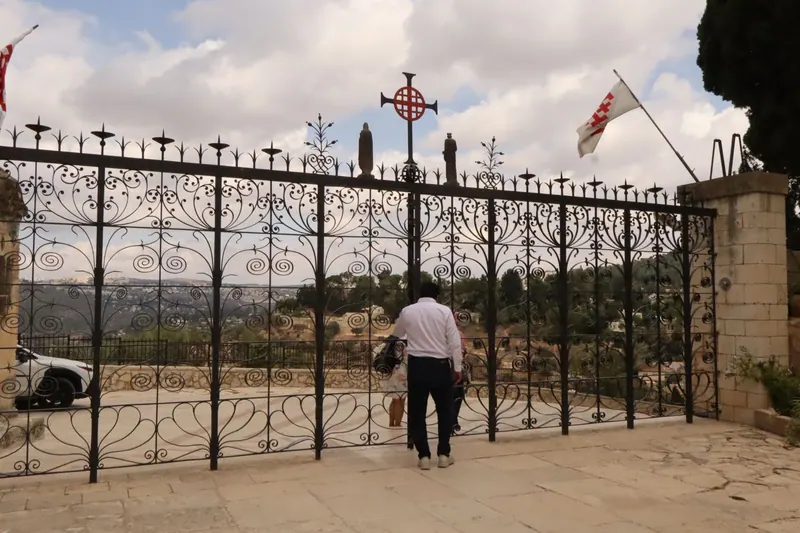 שער ברזל מסולסל ובראשו צלב ירושלים אדום ושני פסלונים, דגלים עם הצלב מתנופפים; מבקר מביט דרכו אל הגבעות המיוערות.