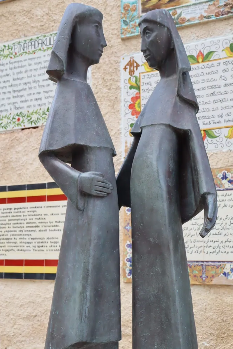 פסל ברונזה של מרים ואלישבע עומדות זו מול זו, ומאחוריהן לוחות אריחים כתובים בשפות רבות.