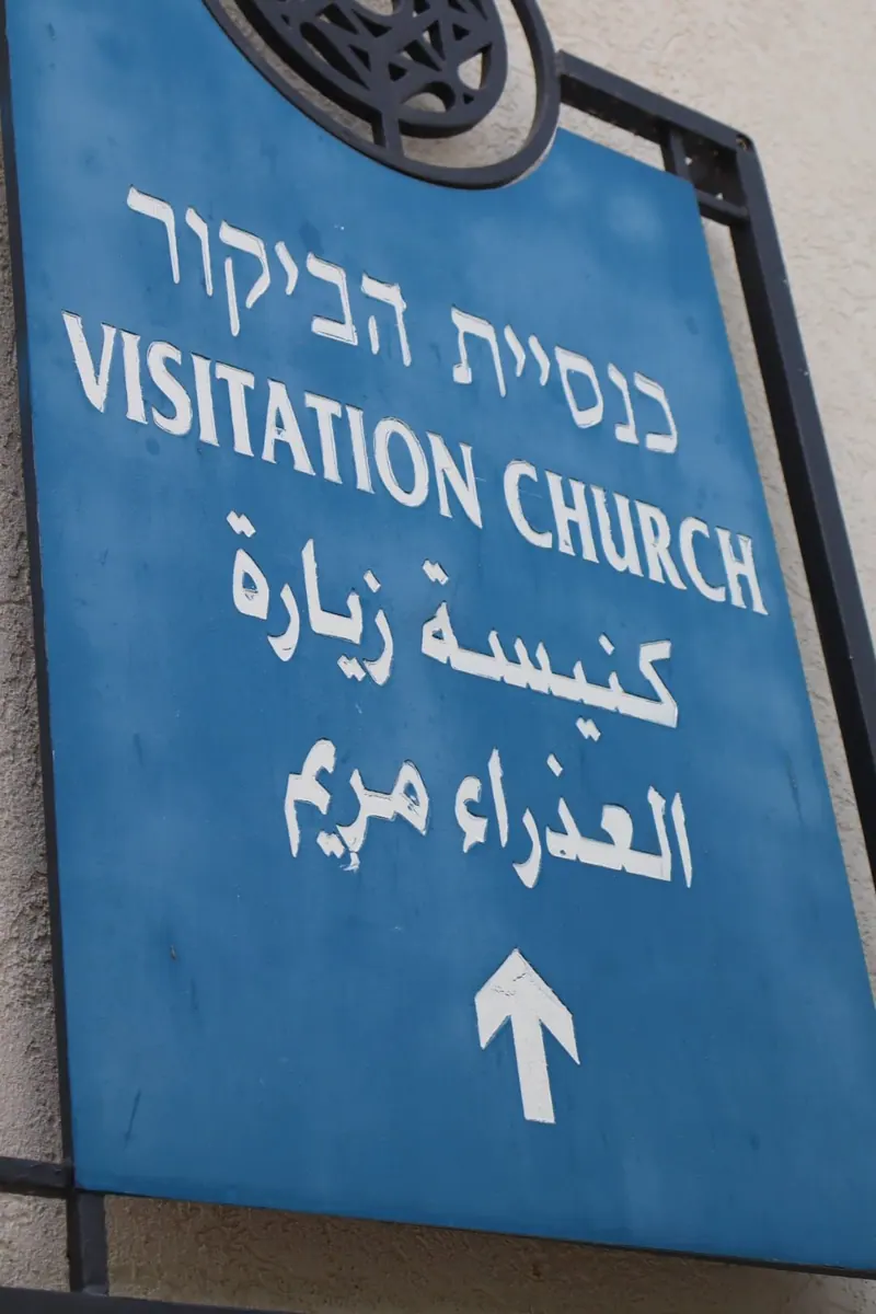 שלט כחול "כנסיית הביקור" בעברית, באנגלית ובערבית, עם חץ כלפי מעלה.