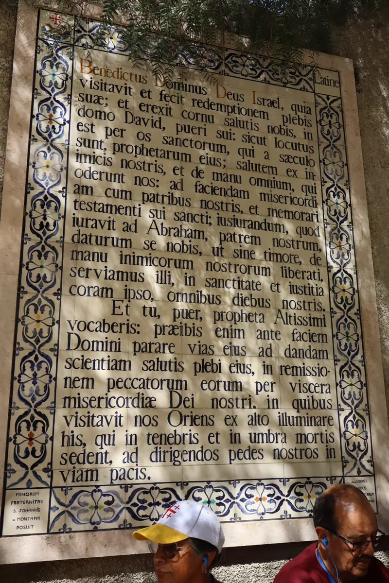 לוח אריחים בלטינית עם תפילת Benedictus ומסגרת עיטורים כחולה; בתחתיתו שני מבקרים, אחד בכובע מצליה.