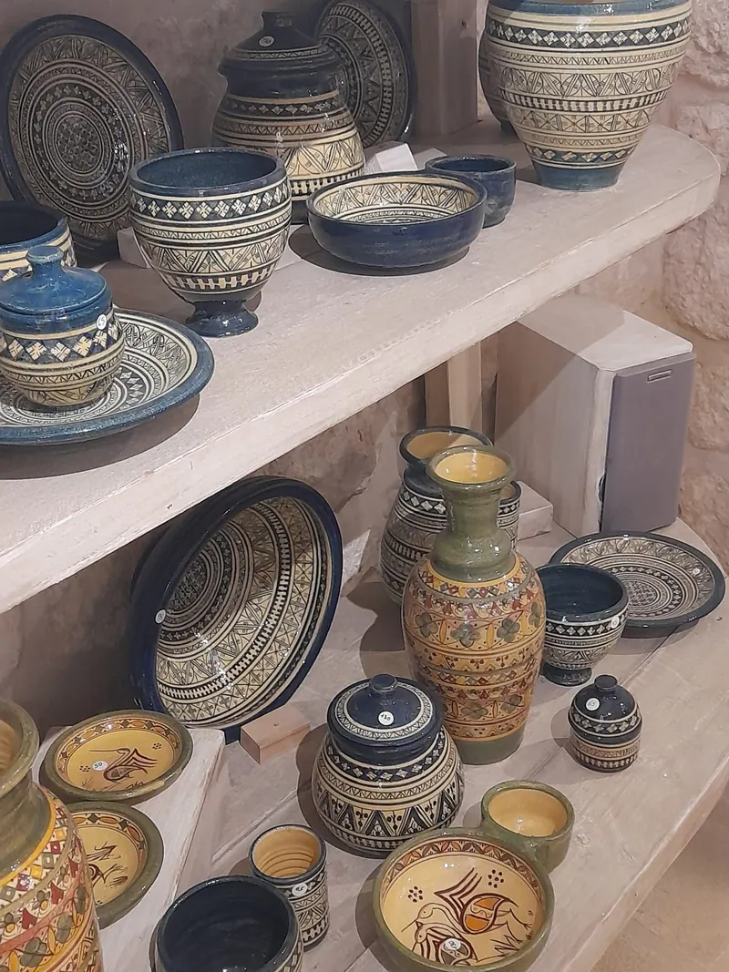 מדפים של כלי קרמיקה מצוירים ביד: קערות, צלחות וכדים בכחול, צהוב וירוק עם דוגמאות גאומטריות וציפורים.