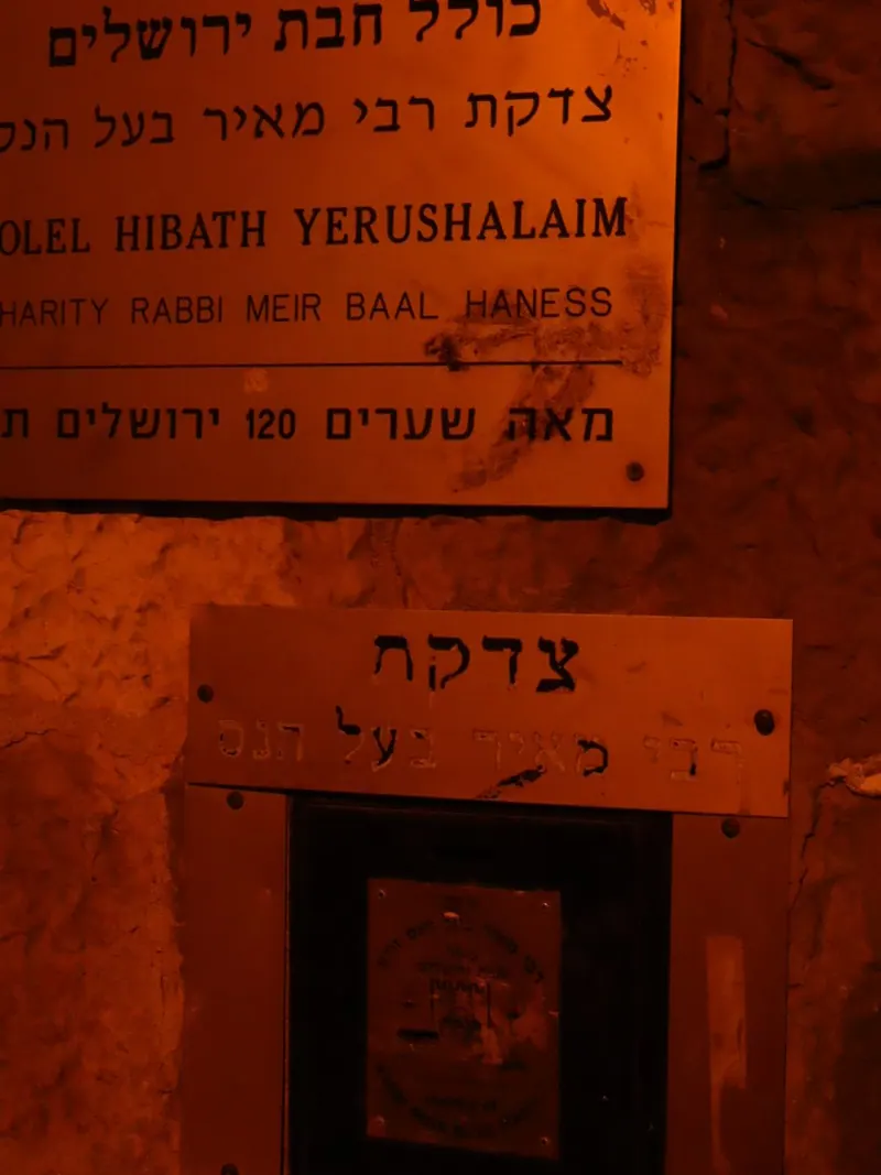 שלט "כולל חבת ירושלים – צדקת רבי מאיר בעל הנס, מאה שערים 120" ומתחתיו קופת צדקה בקיר.