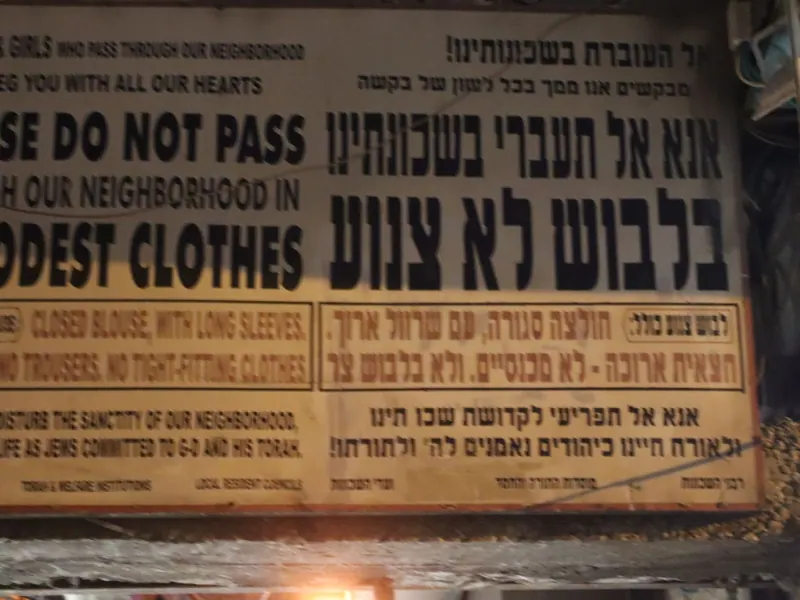 שלט דו-לשוני בעברית ובאנגלית: "אנא אל תעברי בשכונתינו בלבוש לא צנוע", עם פירוט הלבוש הנדרש.
