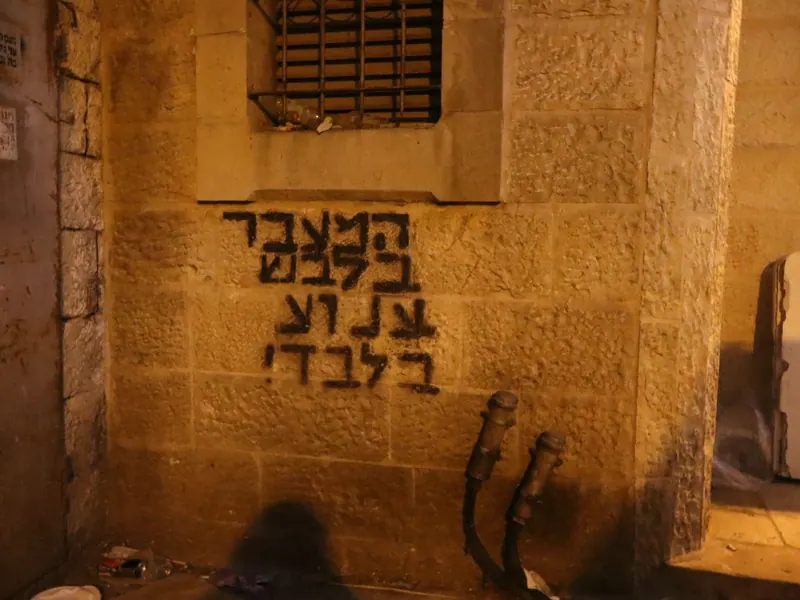 כתובת מרוססת בשחור על קיר אבן מואר בלילה: "המעבר בלבוש צנוע בלבד!", מתחת לחלון מסורג.