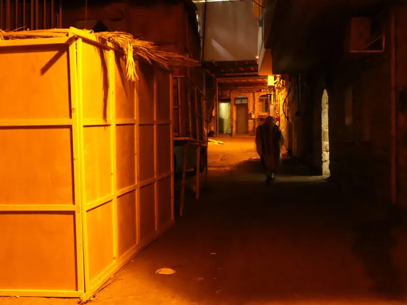 סמטה מוארת בכתום בלילה: סוכת עץ עם סכך כפות תמרים, ובהמשך מתפלל עטוף טלית צועד.