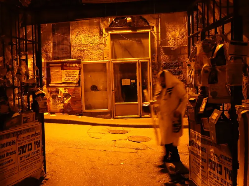 מעבר בלילה באור כתום, שעריו מכוסים מודעות "אין כניסה למאה שערים", ומתפלל עטוף טלית חולף בו.