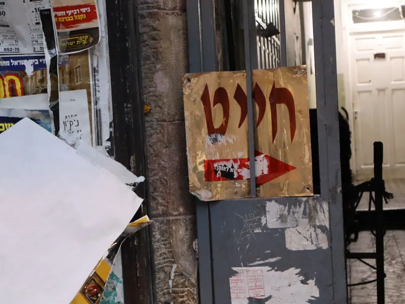 שלט פח ועליו "חייט" וחץ אדום, תלוי על שער ברזל אפור ליד מודעות קרועות.