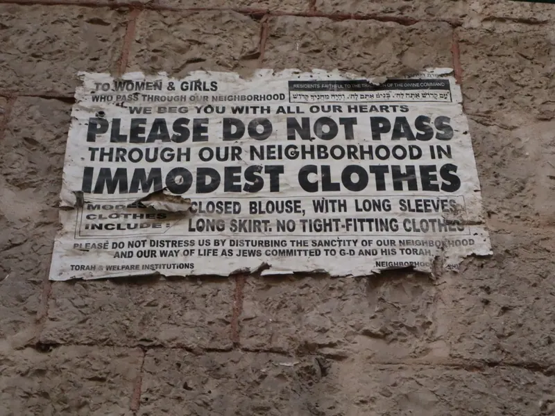 שלט מודפס וקרוע על קיר אבן, באנגלית: "PLEASE DO NOT PASS THROUGH OUR NEIGHBORHOOD IN IMMODEST CLOTHES".