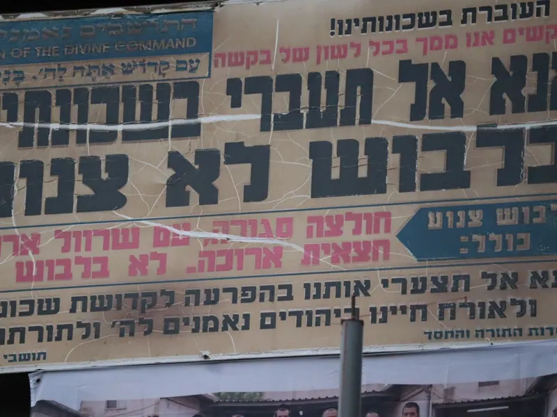 שלט סדוק וישן: "אנא אל תעברי בשכונתינו בלבוש לא צנוע", ובו פירוט באדום של חולצה סגורה וחצאית ארוכה.