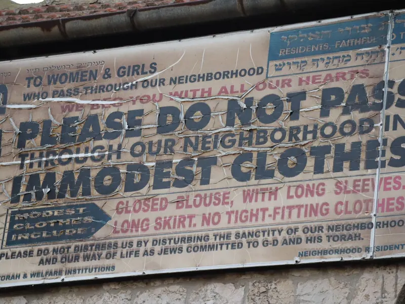 שלט סדוק באנגלית: "TO WOMEN & GIRLS... PLEASE DO NOT PASS THROUGH OUR NEIGHBORHOOD IN IMMODEST CLOTHES".