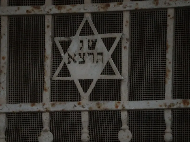 מגן דוד מברזל צבוע לבן בתוך סורג חלון חלוד, ובמרכזו אותיות עבריות.