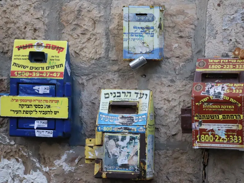 ארבע קופות צדקה מפח צבעוני תלויות על קיר אבן, עליהן שמות מוסדות ומספרי טלפון.