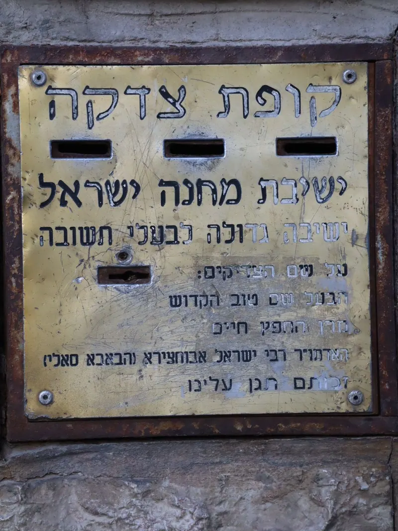 לוח פליז שחוק עם חריצים לכסף ועליו הכיתוב "קופת צדקה – ישיבת מחנה ישראל" ושמות צדיקים.
