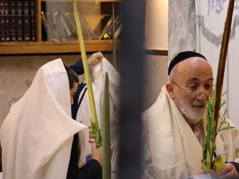 מתפלל מבוגר עטוף טלית מחזיק לולב ואתרוג, ולידו נער שטליתו מכסה את ראשו, ליד מדף ספרי קודש.