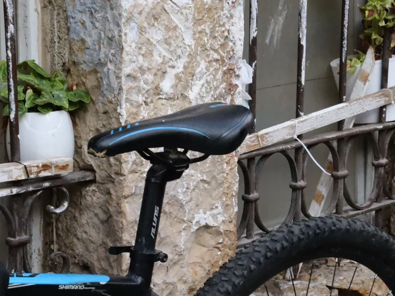 מושב אופניים שחור ליד קיר אבן מתקלף, מעקה ברזל מעוטר ועציץ לבן על אדן חלון.