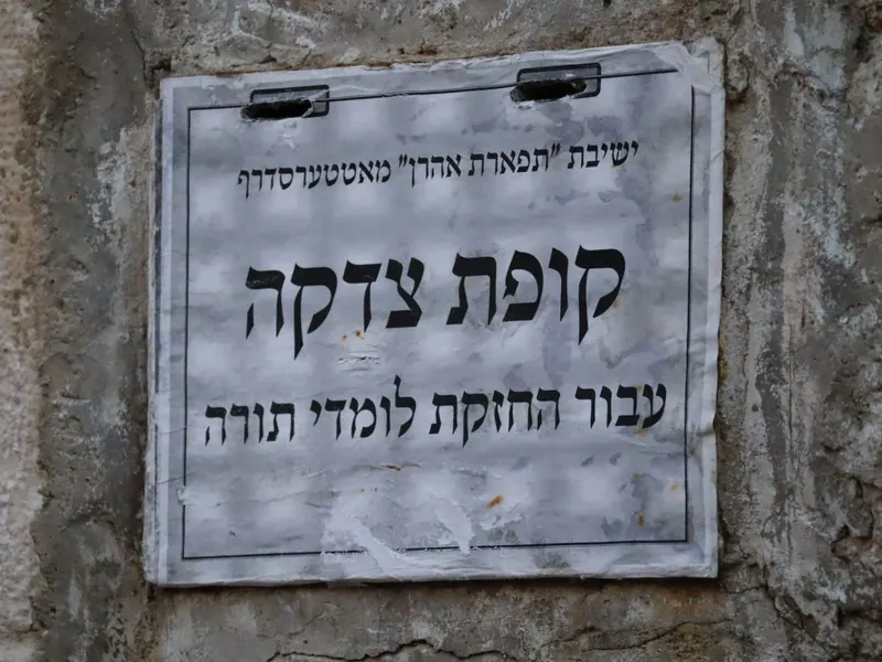 לוח מתכת לבן על קיר: "קופת צדקה עבור החזקת לומדי תורה" של ישיבה.