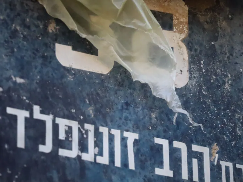 שלט רחוב כחול ישן ומתקלף, "רח' הרב זוננפלד", ושקית ניילון תלויה עליו.