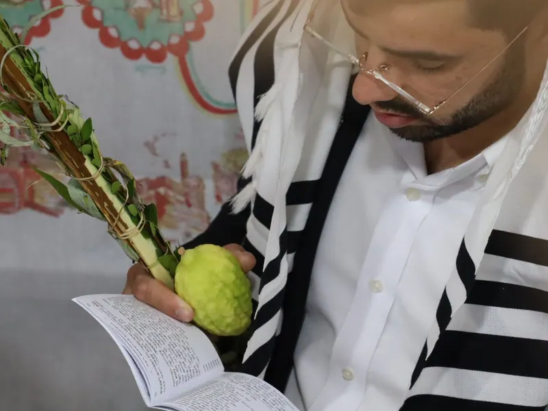 מתפלל עטוף טלית אוחז לולב ואתרוג וקורא בסידור, ומאחוריו קישוט בד צבעוני של סוכה.