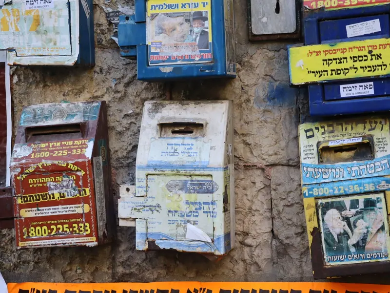 קופות צדקה ישנות מפח צבעוני תלויות על קיר אבן, מכוסות מדבקות, כיתובים ותמונות רבנים.