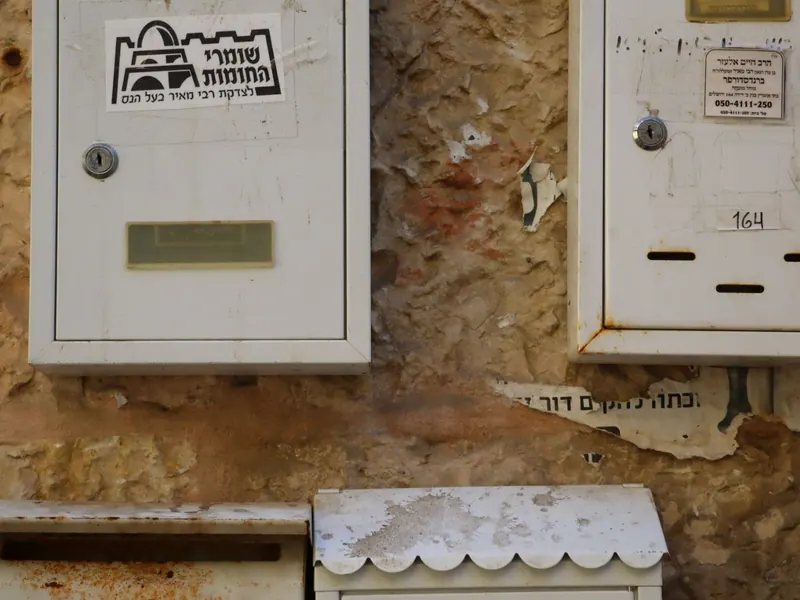 תיבות דואר מתכת דהויות על קיר אבן מתקלף, ועל אחת מדבקה "שומרי החומות – לצדקת רבי מאיר בעל הנס".