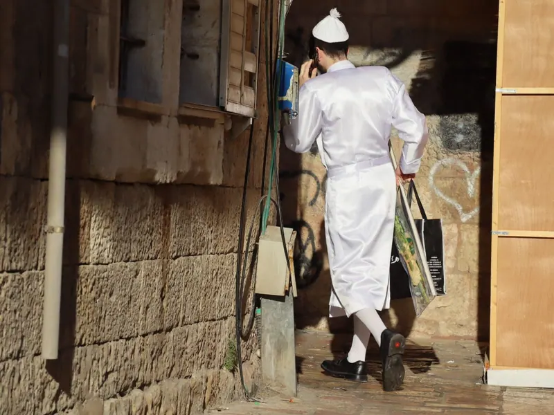 גבר בגלימה לבנה מבריקה, גרביים לבנות וכיפה לבנה מדבר בטלפון ונושא שקית עם לולב, ליד קיר אבן.