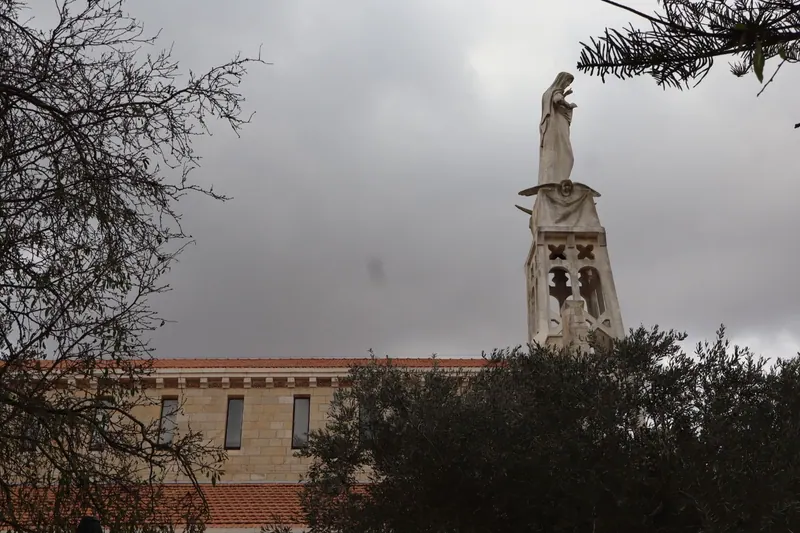 פסל דמות בגלימה ניצב על ראש מגדל אבן מגולף, מעל גג רעפים אדום ועצי זית, תחת שמיים אפורים.