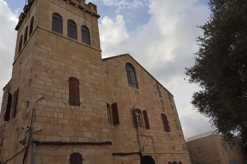 בניין אבן גבוה עם מגדל משונן, חלונות קשתיים ותריסי עץ חומים, ולצדו עץ ירוק תחת שמיים מעוננים.