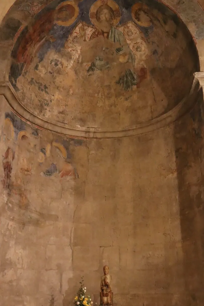 ציור קיר דהוי בכיפת אפסיס: דמות מוכתרת בהילה במרכז ודמויות במדליונים סביבה; למטה פסלון עץ של אם וילד וזר פרחים.
