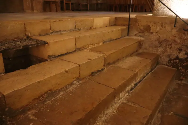 מדרגות אבן עתיקות ושחוקות באור עמום, עם מעקה ברזל ופתח מכוסה סבכה בצד.