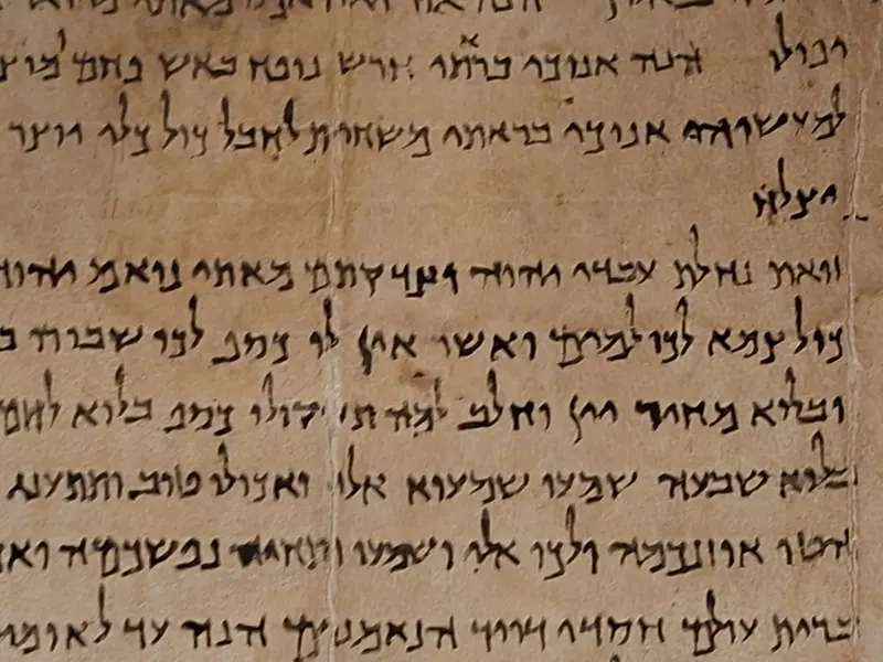 תקריב של קלף עתיק ועליו שורות של כתב עברי בדיו שחורה וסימנים קטנים בשוליים.