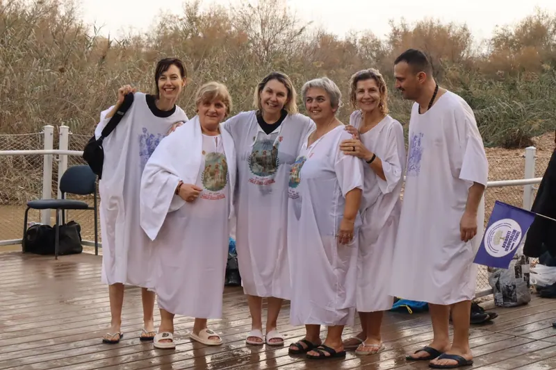 קבוצת מבוגרים בגלימות טבילה לבנות עומדת על רציף עץ על גדת הנהר, בין קני סוף.