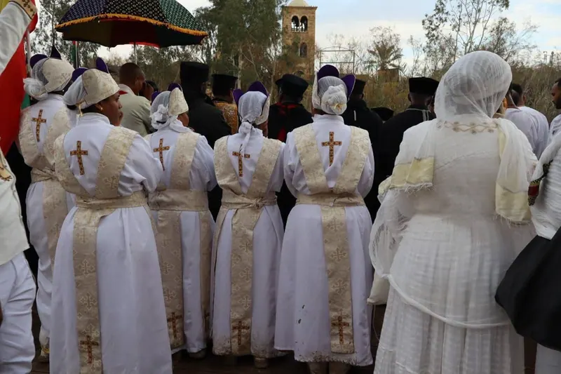 שורת זמרים בגלימות לבנות עם צלבים על הגב מצולמת מאחור, מעליהם שמשייה רקומה, וברקע מגדל כנסייה ועצים.