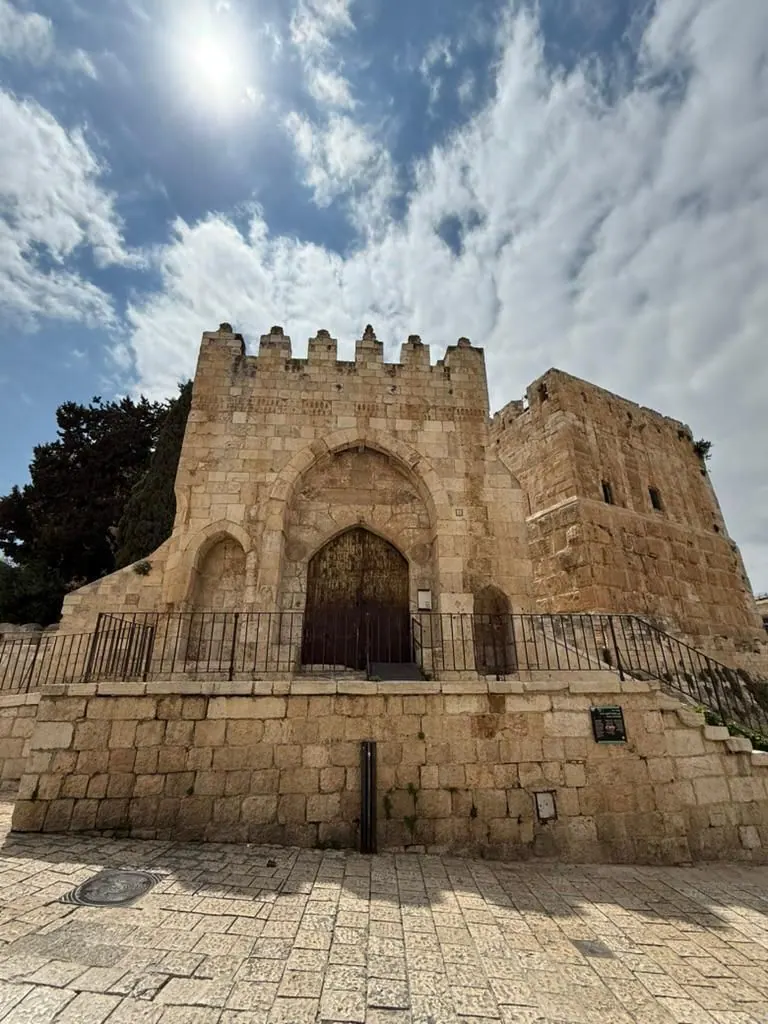 מגדל דוד מבחוץ: חומת אבן ומגדלים עם שער קשתי, ורחבה מרוצפת לפניהם תחת שמיים כחולים.